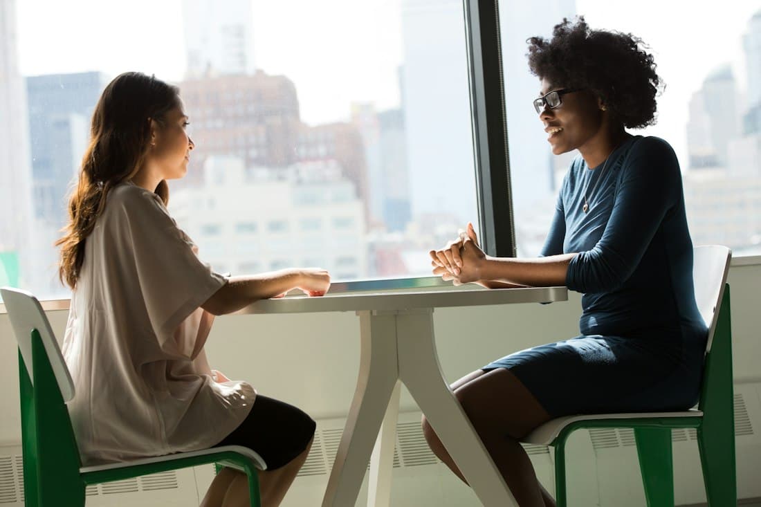 Two women sitting across a small white table by a tall window with city buildings behind them, talking